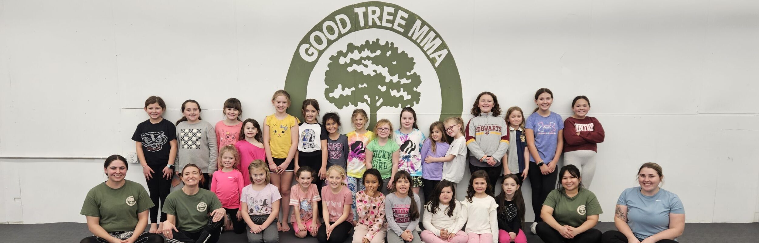 kids' class members stand in front of the Good Tree MMA Logo on our gym wall
